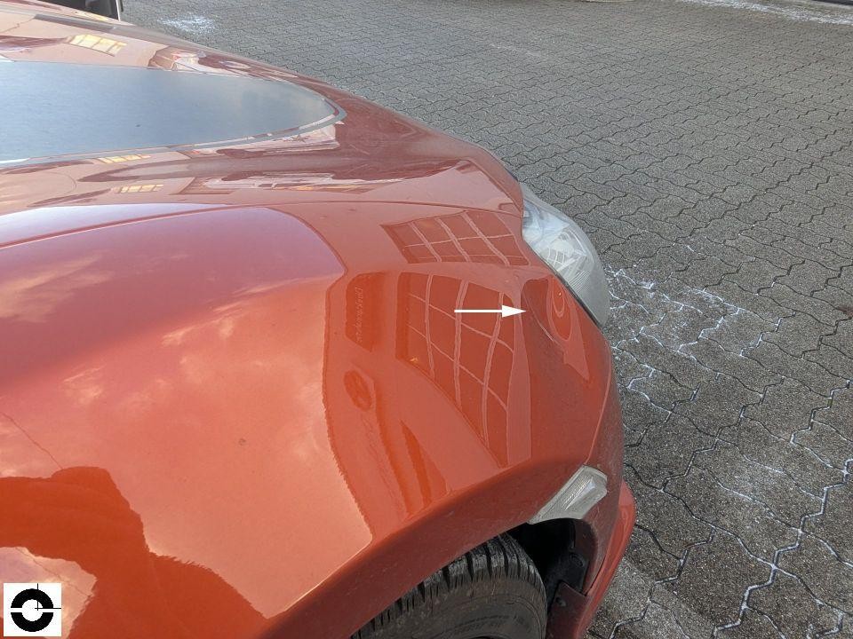 Frontansicht, Motorhaube mit weißem Pfeil markiert Delle/Verformung, Scheinwerfer rechts sichtbar