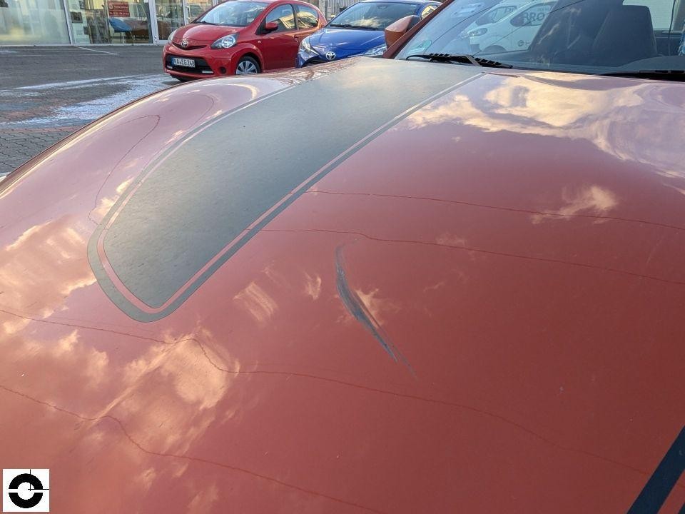 Motorhaube von oben, Kratzer und Lackschäden auf orangefarbener Oberfläche mit grauem Streifen